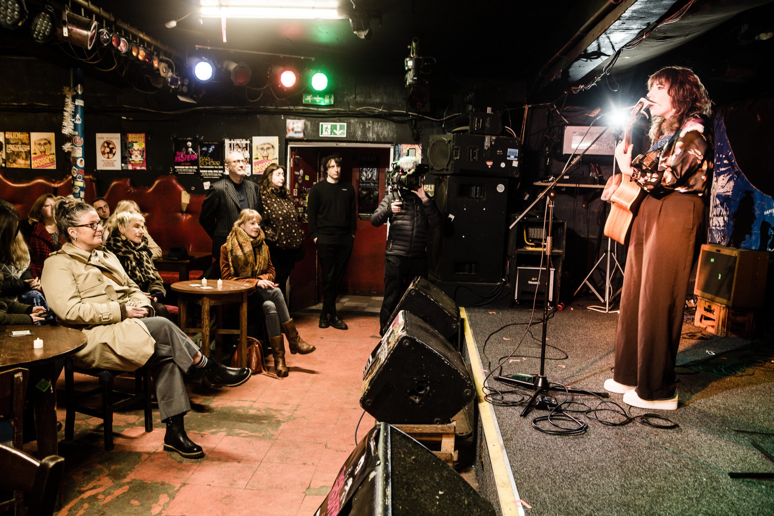 A female singer performs on a stage with a guitar in a dark venue with an audience watching