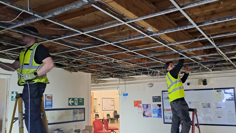 Two people wearing high vis jackets are stood on step ladders rewiring a ceiling in a community centre. Beams are visible and lots of wires hang down. Stacked chairs and signage can be seen in the far corner through a doorway.