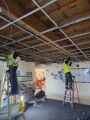 Two people wearing high vis jackets are stood on step ladders rewiring a ceiling in a community centre. Beams are visible and lots of wires hang down. Stacked chairs and signage can be seen in the far corner through a doorway.