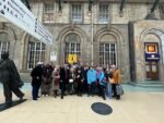 A mixed group of 30 people stood in front of a cream brick building with a statue to the left and train station signage above