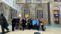 A mixed group of 30 people stood in front of a cream brick building with a statue to the left and train station signage above