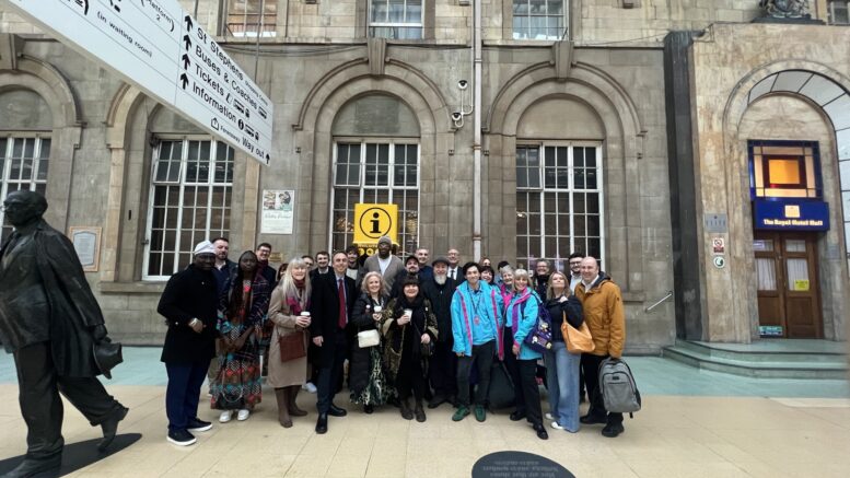 A mixed group of 30 people stood in front of a cream brick building with a statue to the left and train station signage above