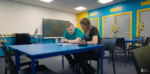 A young man sat at a blue table with his mentor during his Supported Internship. They are both reading and writing on a document together.