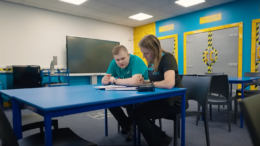 A young man sat at a blue table with his mentor during his Supported Internship. They are both reading and writing on a document together.