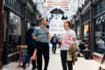 2 young men appear to be walking through a covered shopping arcade with bunting hanging from the ceiling. Both have a disposable coffee cup in hand.