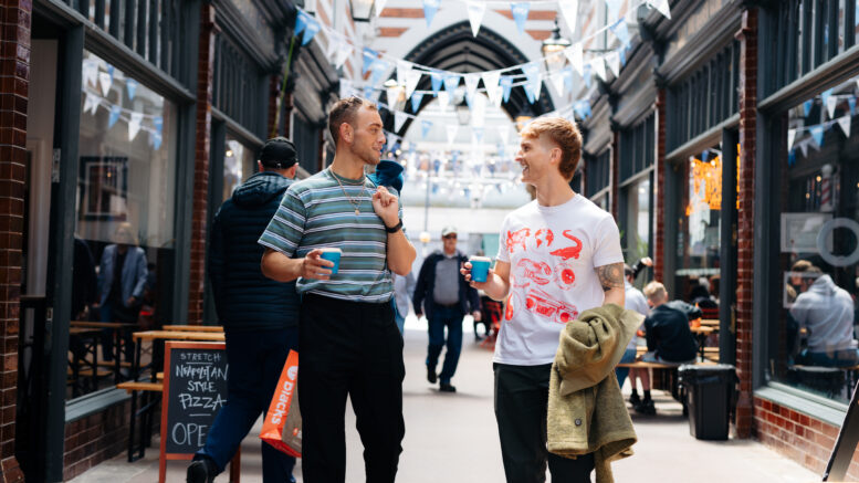 2 young men appear to be walking through a covered shopping arcade with bunting hanging from the ceiling. Both have a disposable coffee cup in hand.