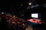 A wide shot of a theatre style space with people sat in red seats facing a red stage with people sat on the stage and a large screen behind them