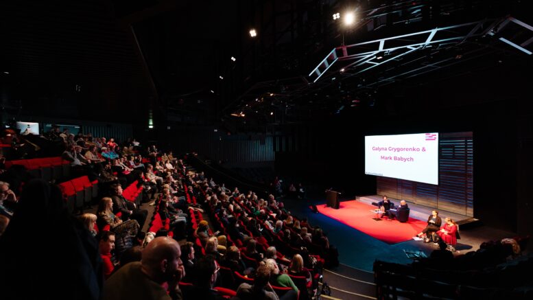 A wide shot of a theatre style space with people sat in red seats facing a red stage with people sat on the stage and a large screen behind them