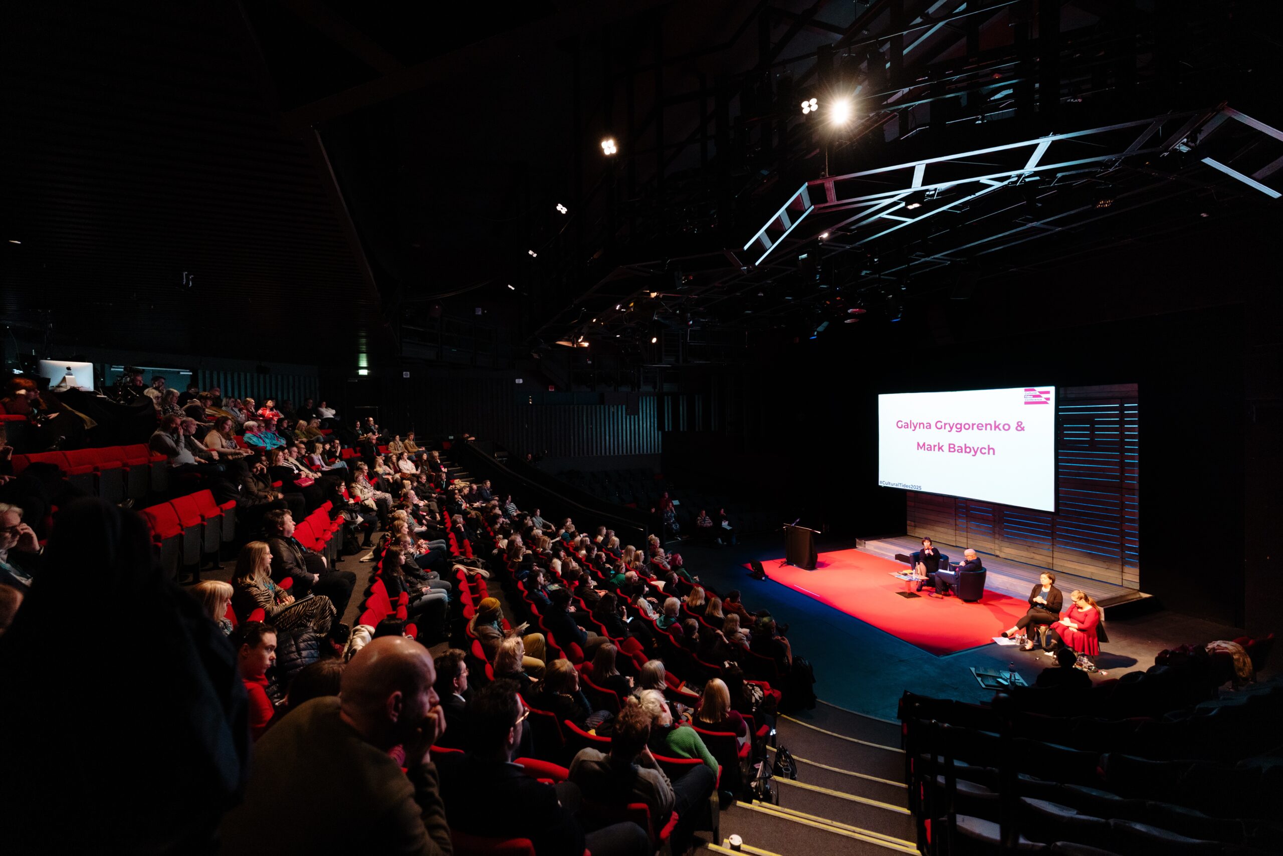 A wide shot of a theatre style space with people sat in red seats facing a red stage with people sat on the stage and a large screen behind them