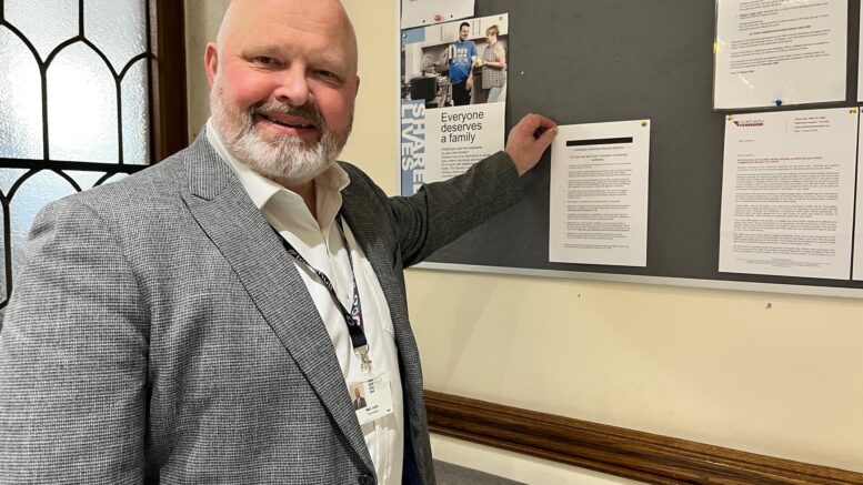 A man in a grey suit pins a piece of paper to a grey notice board