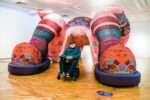 A man with a flat cap sits in a wheelchair in front of a large inflatable pair of legs