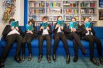 A group of school children in uniform sit on a sofa all with the same book open in front of her face.