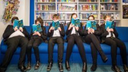 A group of school children in uniform sit on a sofa all with the same book open in front of her face.