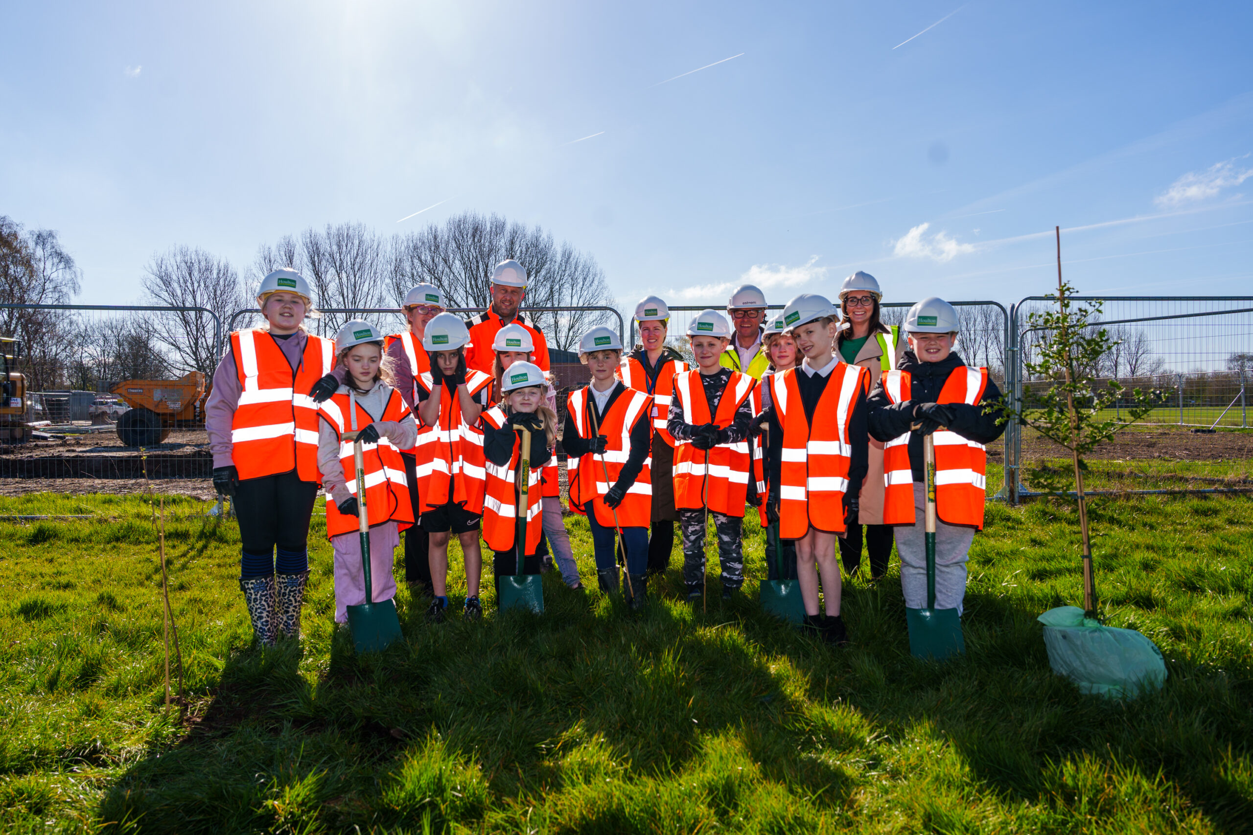 a group of children and adults stand together on a field with orange high vis bibs and hard hats