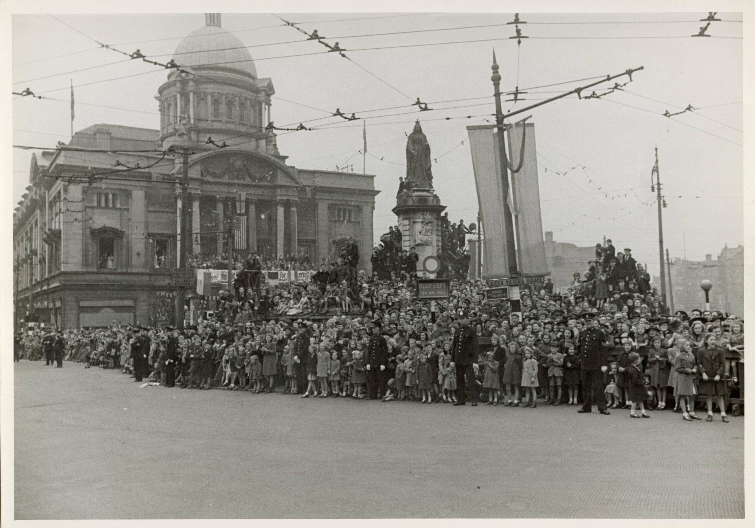 An old black and white image of Queen Victoria Square in Hull with crowds of people sat on the monument and City Hall in the background