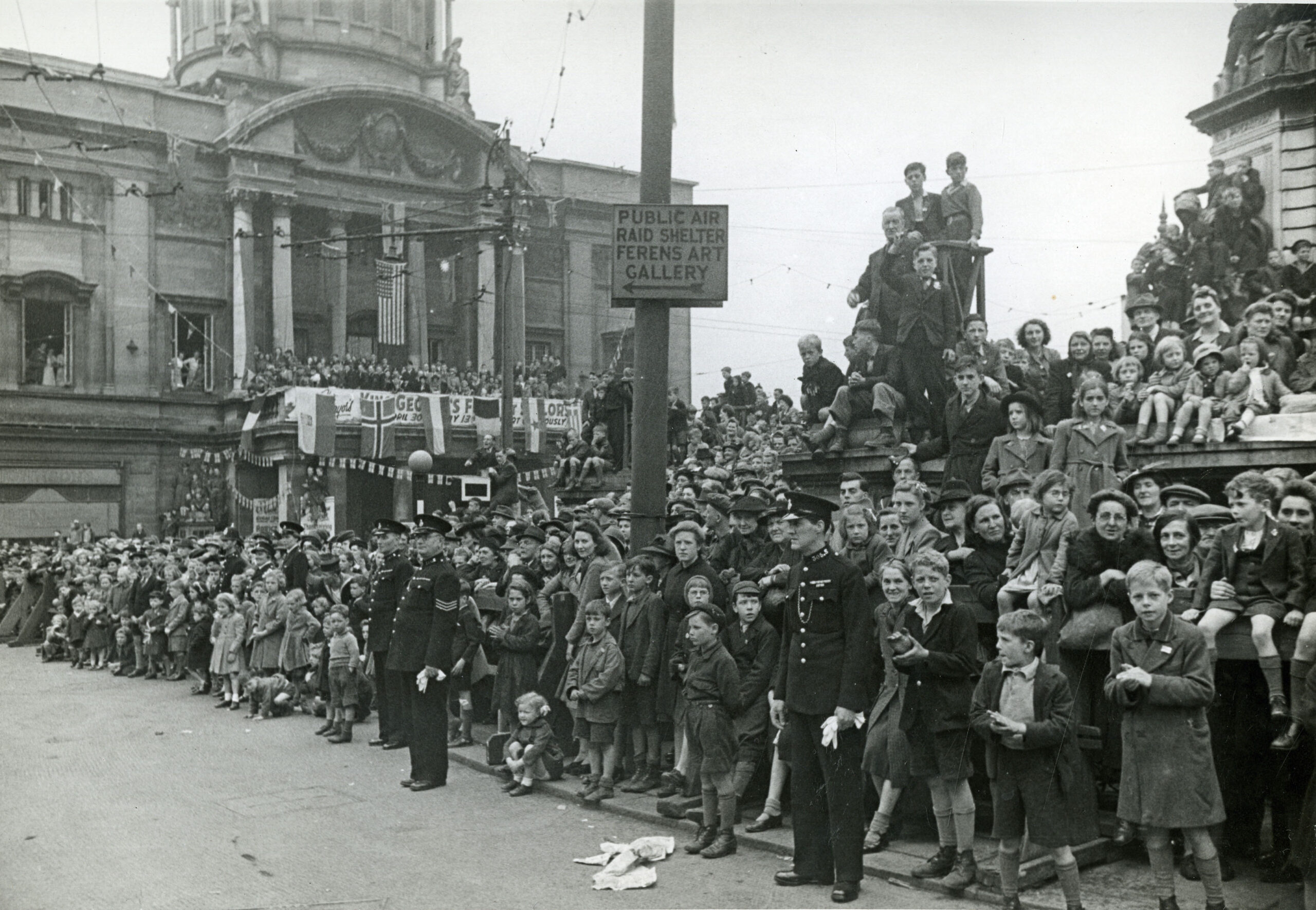 The photo is black and white. It shows crowds of mostly women and children lined up in Hull city centre, ready to watch the victory parade. The city hall in the background is adorned with the flags of the allies.