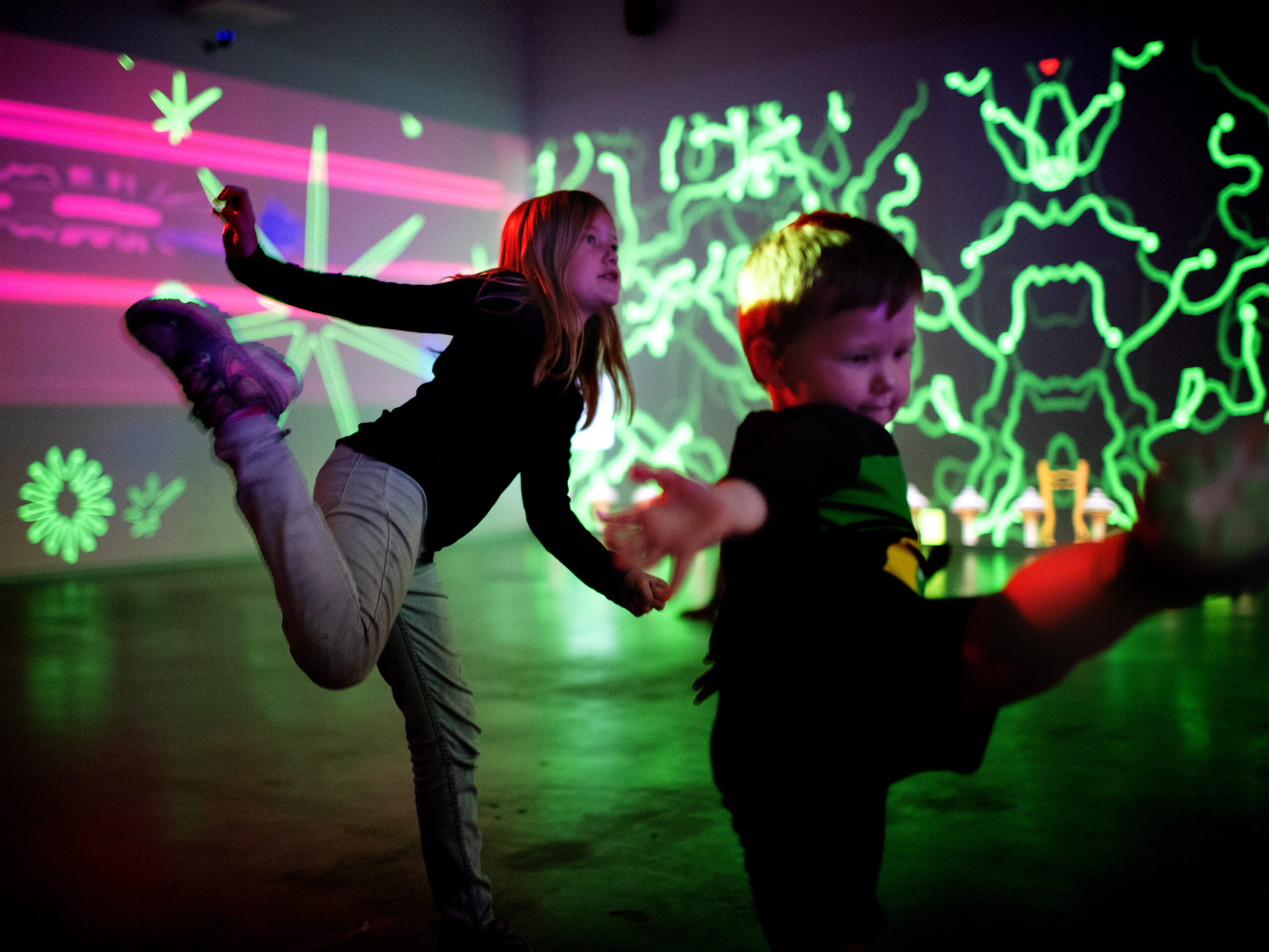 a young girl and boy appear to be dancing in a large dark room with neon graffiti style drawing on the walls behind them