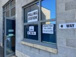 A window in a grey building with a polling station poster and arrows on it pointing to an open door