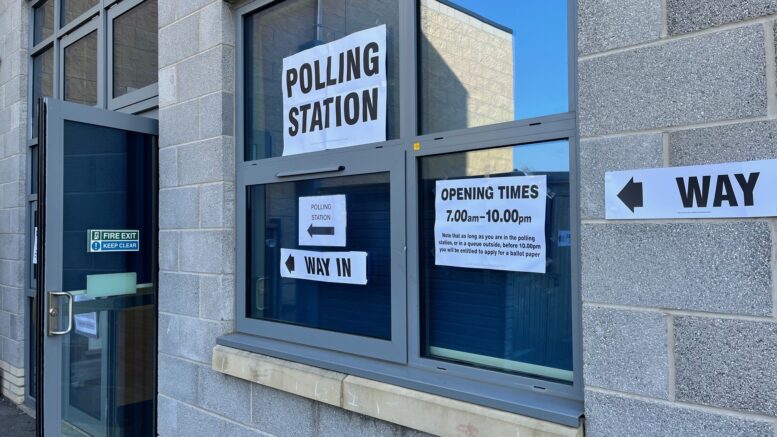 A window in a grey building with a polling station poster and arrows on it pointing to an open door