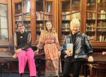 3 woman pose together on dark wooden tables in front of glass bookcases filled with books