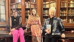 3 woman pose together on dark wooden tables in front of glass bookcases filled with books