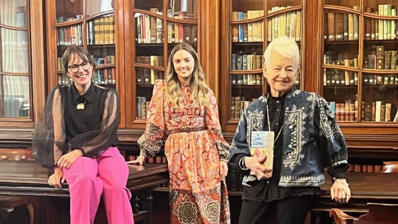 3 woman pose together on dark wooden tables in front of glass bookcases filled with books