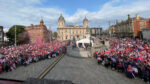 A wide elevated shot in an open city square with light stone buildings in the background and thousands of people dressed in red and white in the foreground