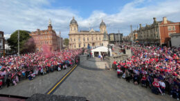 A wide elevated shot in an open city square with light stone buildings in the background and thousands of people dressed in red and white in the foreground