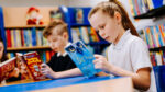 a girl and a boy sit at a table reading books, with shelves of books in the background