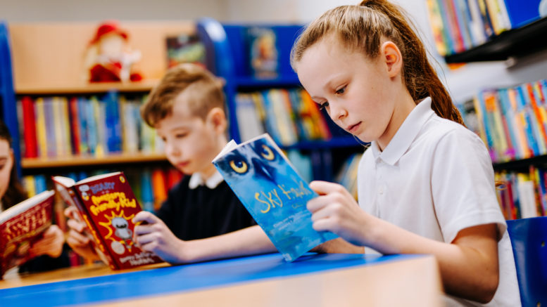 a girl and a boy sit at a table reading books, with shelves of books in the background