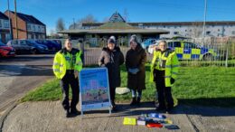 Two uniformed police officers and two council officers stand with an A-board and some giveaways. A police car can be seen in the background