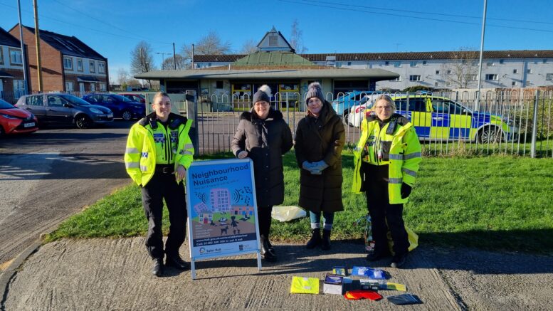 Two uniformed police officers and two council officers stand with an A-board and some giveaways. A police car can be seen in the background