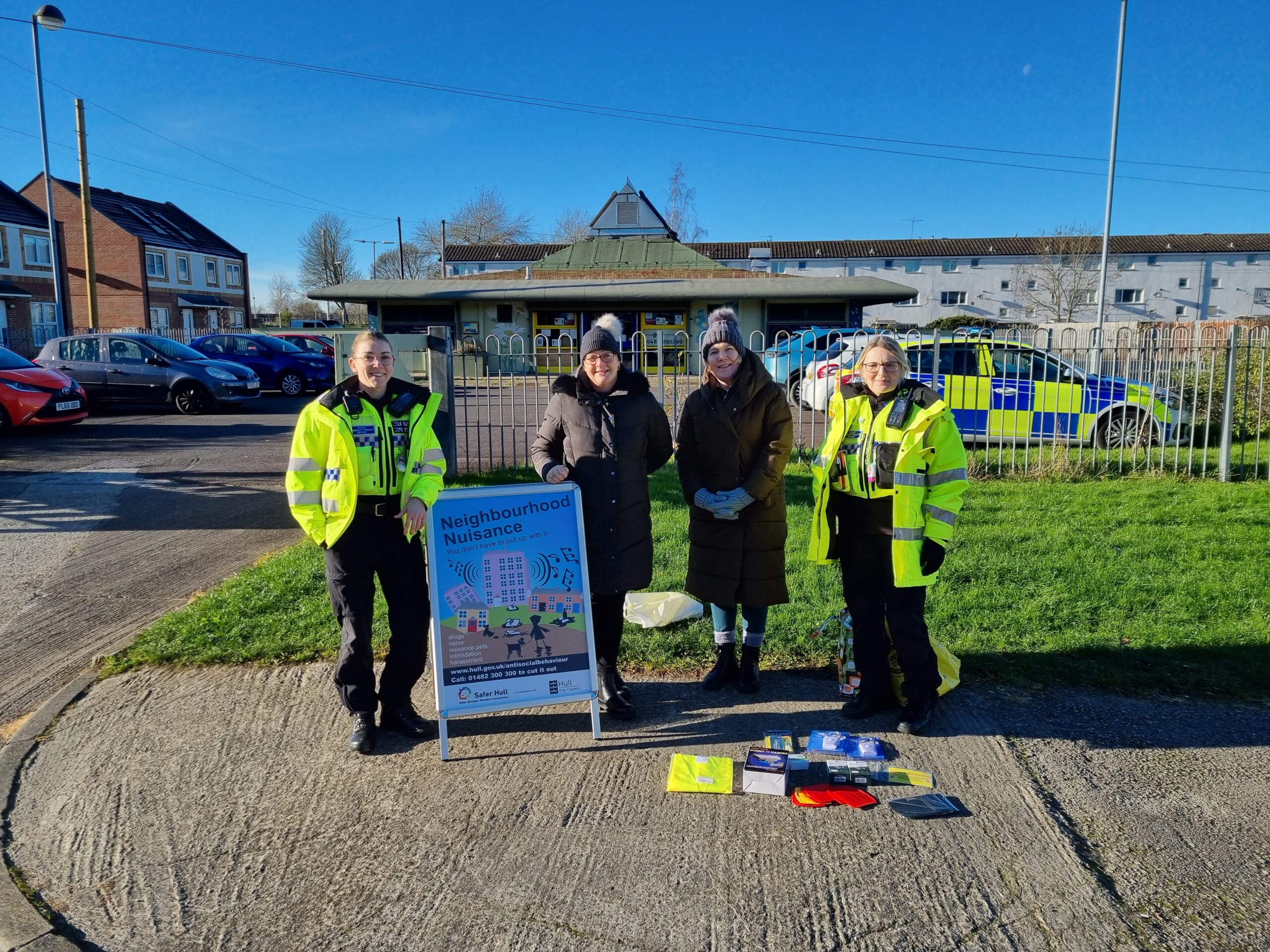 Two uniformed police officers and two council officers stand with an A-board and some giveaways. A police car can be seen in the background