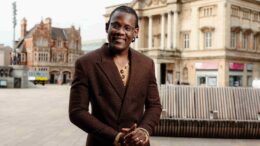 A man in a brown suit stands in the foreground with Hull City Hall behind him