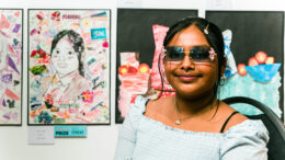 A girl with sunglasses, black hair and a blue dress sits next to a portrait she has drawn.