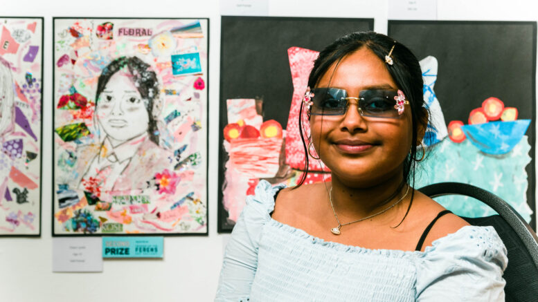 A girl with sunglasses, black hair and a blue dress sits next to a portrait she has drawn.