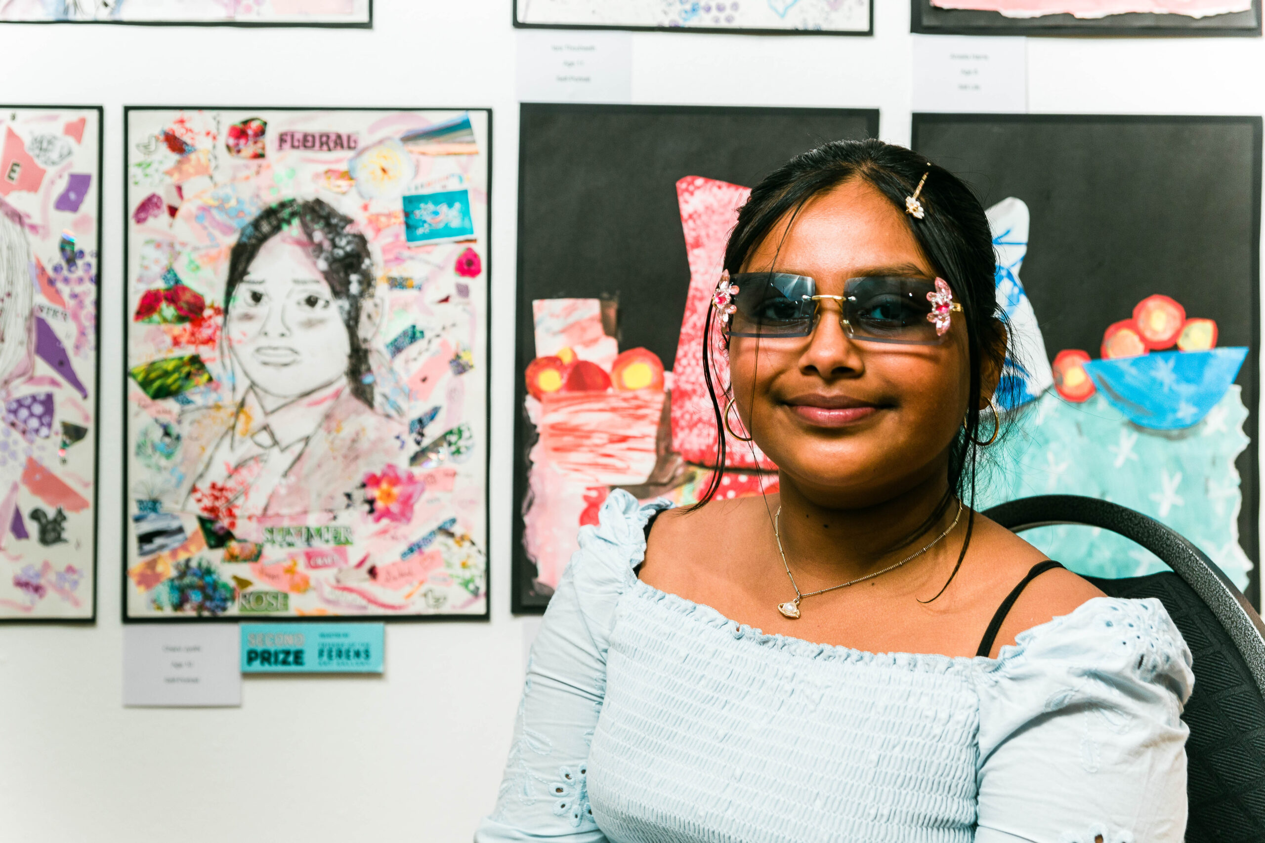 A girl with sunglasses, black hair and a blue dress sits next to a portrait she has drawn.