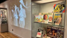 In the foreground a glass cabinet with 1970s memorabilia including board games. Behind a large projection of a black and white image of 2 men with monkeys on their shoulders