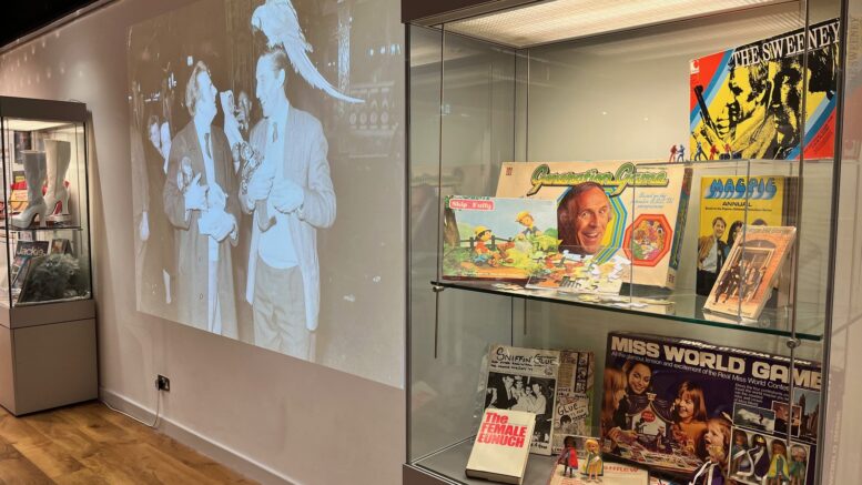 In the foreground a glass cabinet with 1970s memorabilia including board games. Behind a large projection of a black and white image of 2 men with monkeys on their shoulders
