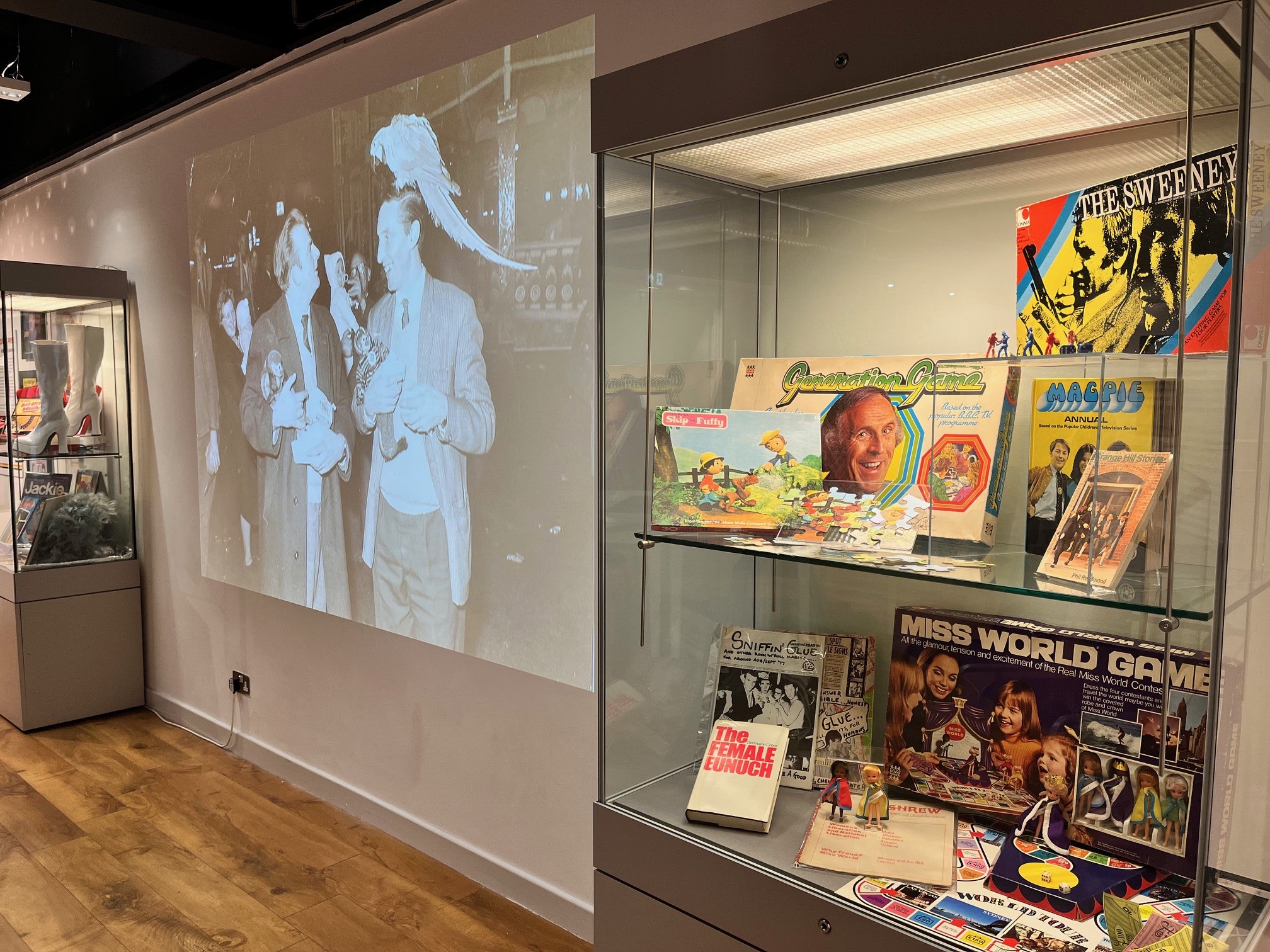In the foreground a glass cabinet with 1970s memorabilia including board games. Behind a large projection of a black and white image of 2 men with monkeys on their shoulders