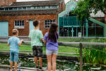 The backs of three young children look over a wooden fence into some gardens with a brick and glass building