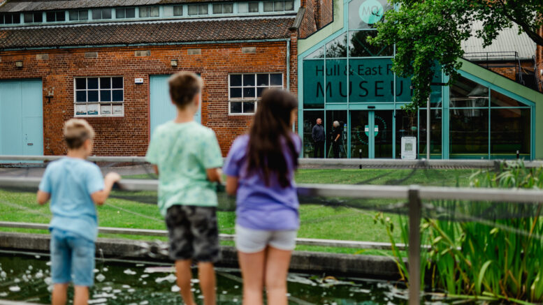 The backs of three young children look over a wooden fence into some gardens with a brick and glass building