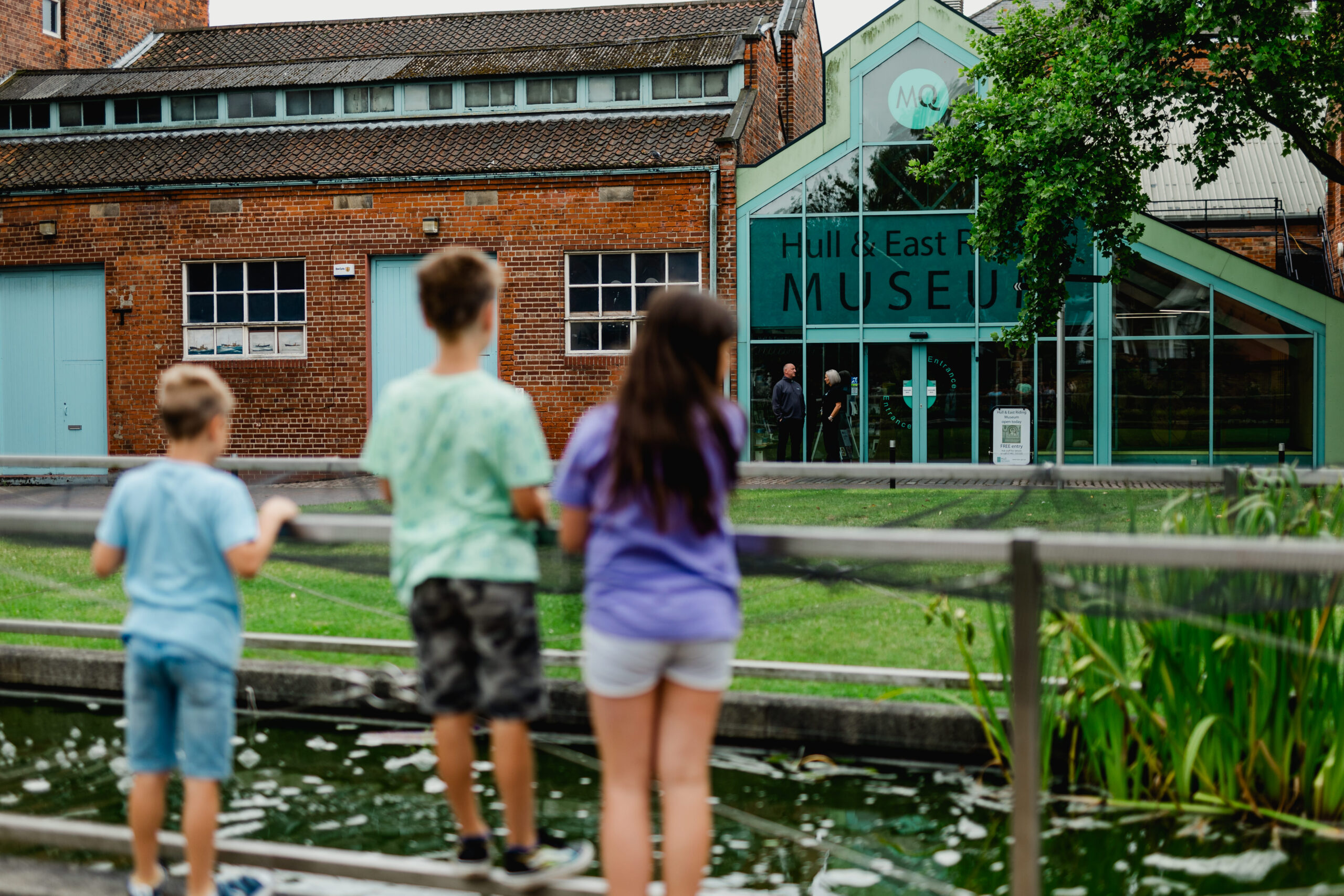 The backs of three young children look over a wooden fence into some gardens with a brick and glass building