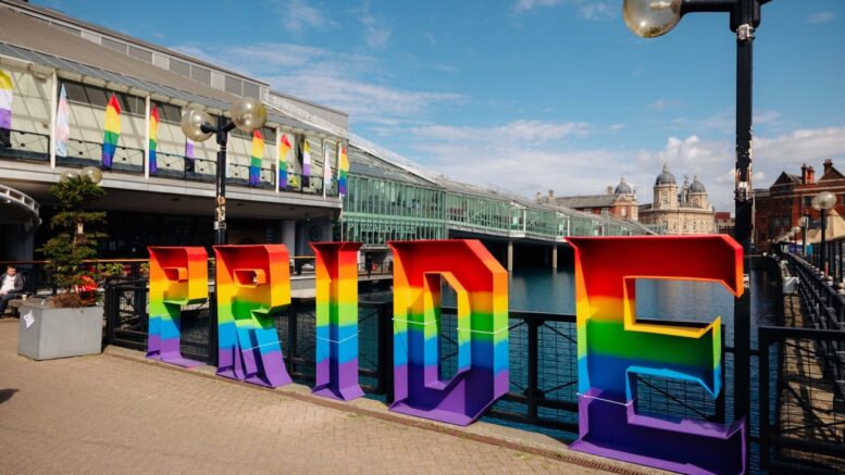 Large letters in rainbow colours spell out PRIDE on a path with large shopping centre and water.