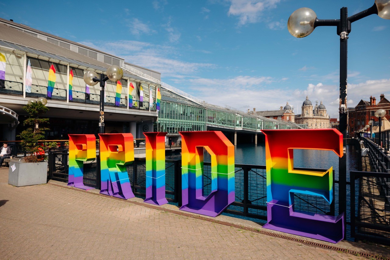 Large letters in rainbow colours spell out PRIDE on a path with large shopping centre and water.