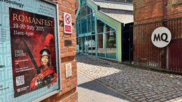 A poster form Romanfest in on an exterior wall in the foreground with the entrance to the Museums Quarter in the background