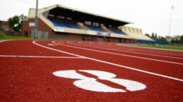 A close up of a red track with a number eight written in white in the forground and a blurred building and seating in the background