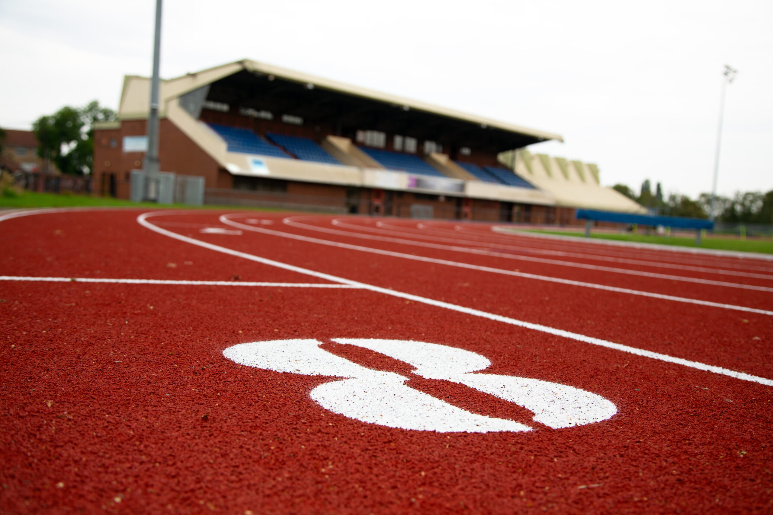 A close up of a red track with a number eight written in white in the forground and a blurred building and seating in the background