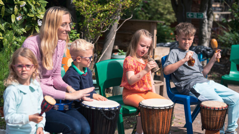 A teacher leads children in an outdoor drumming session in the sun