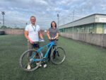 A man and a woman stand behind a blue frame bike on a five-a-side football pitch with a building in the background